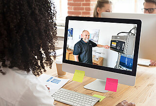 Frau, die vor einem Computer sitzt und jemanden bei der Bedienung einer Maschine beobachtet